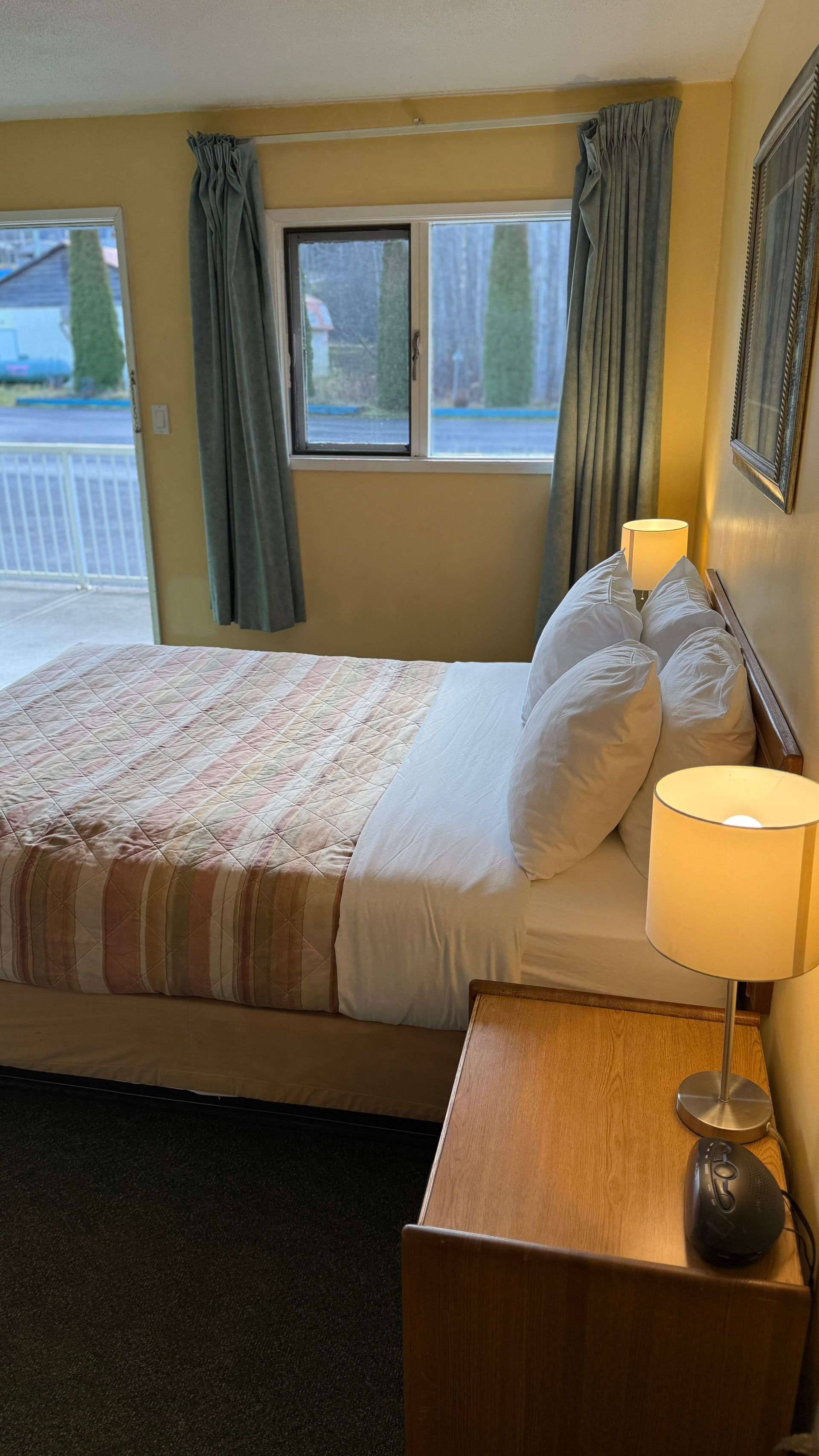 Motel room interior with bed, nightstand, lamps, window, and door to outside area.