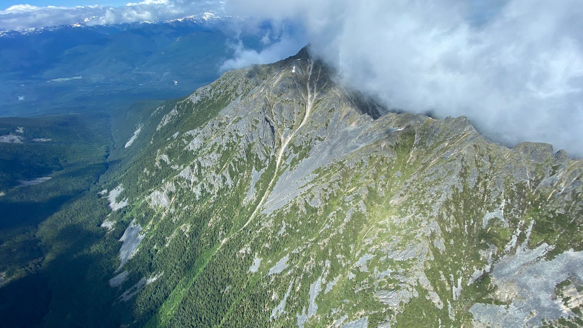 Mountain peak with grey rock, green foliage, and white clouds.