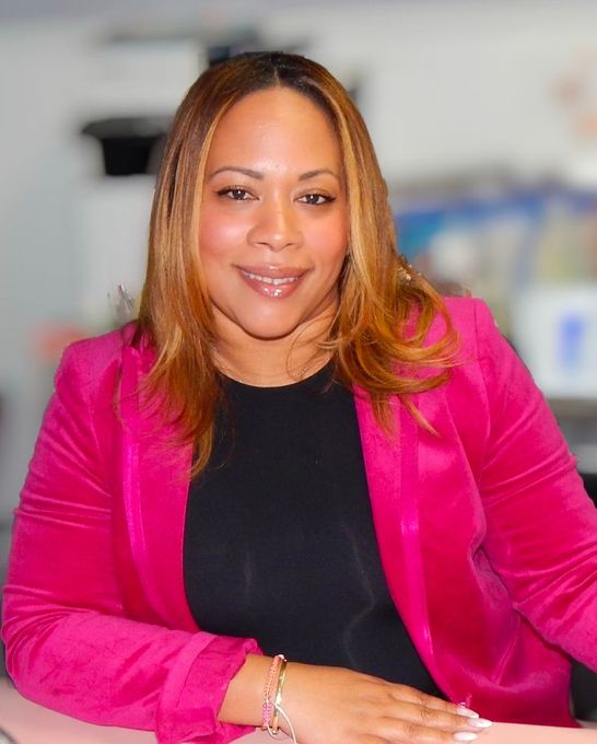 Smiling person in a bright pink blazer and black top, seated against a blurred indoor background