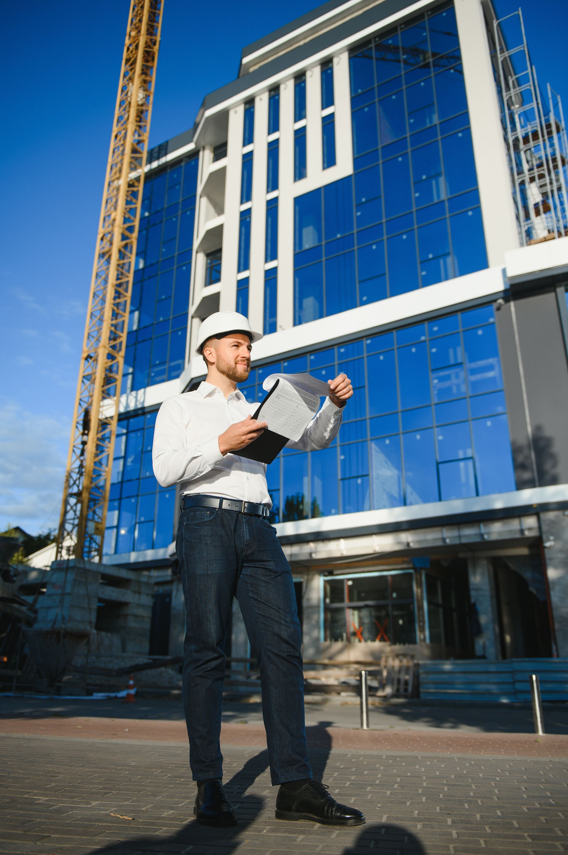 Architect in hard hat reviewing plans outside a modern glass building under construction