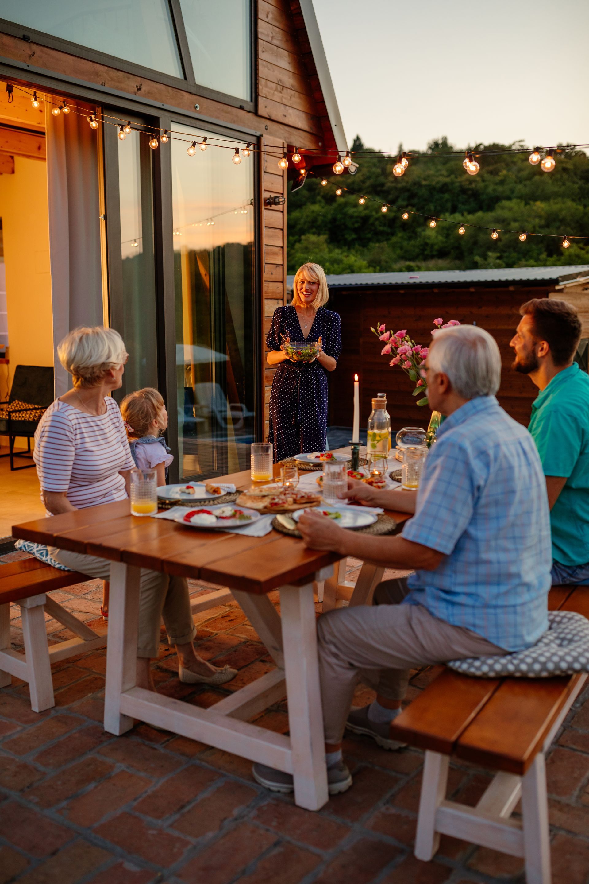 Friends gathered around a wooden table on a patio at sunset, with drinks and food, one person standing and talking