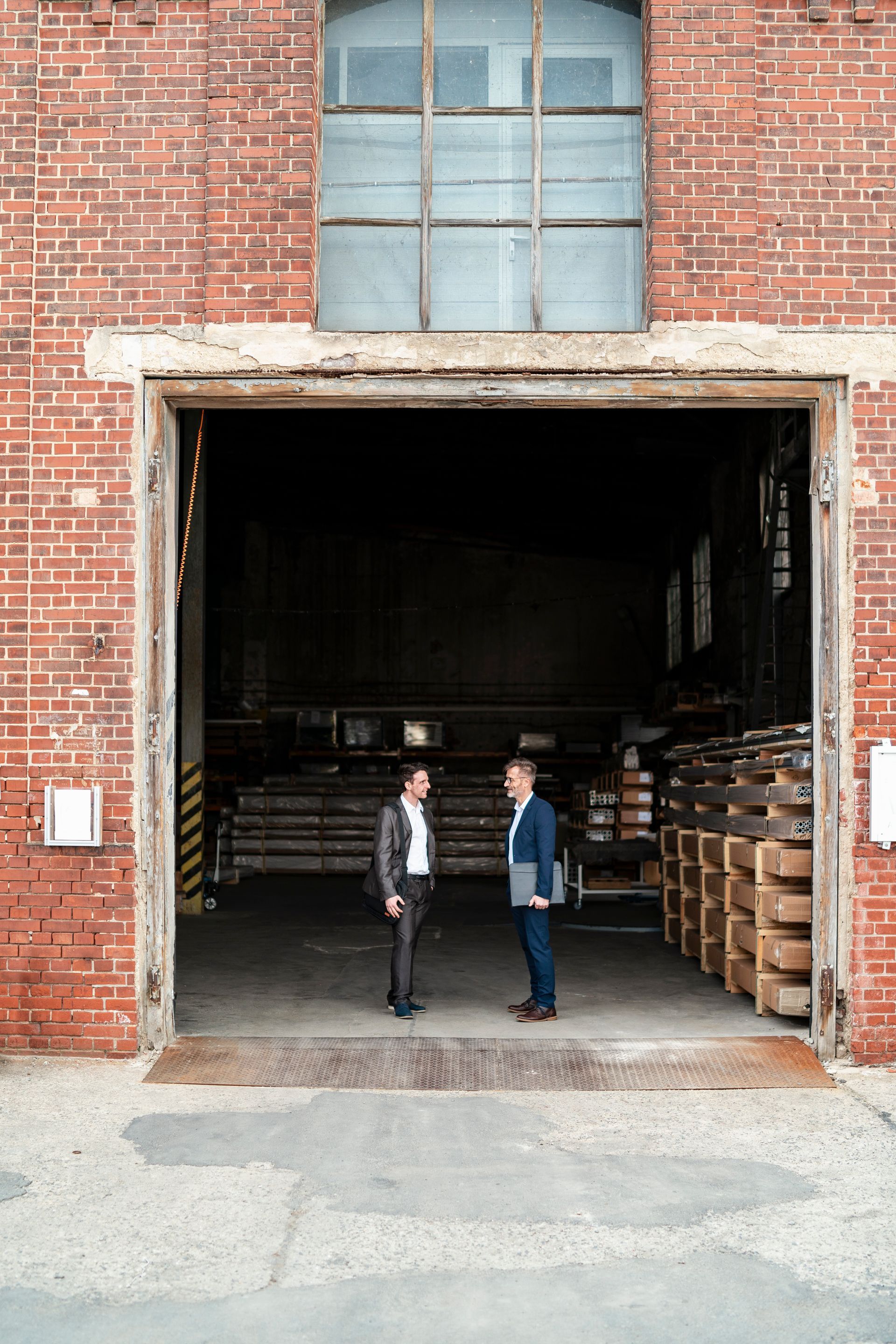 Two people standing in a large brick warehouse doorway, with stacked wooden pallets inside.