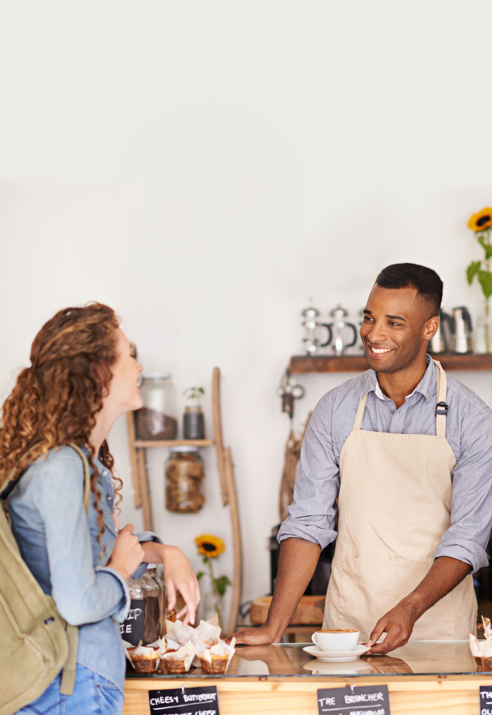 Customer and shopkeeper smiling at a bakery counter, with pastries, cups, and sunflowers in the background.