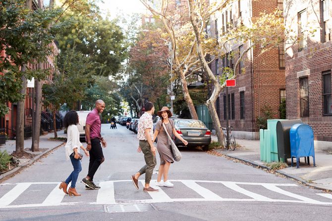 People crossing a tree-lined street at a zebra crossing, with parked cars and brick buildings nearby.