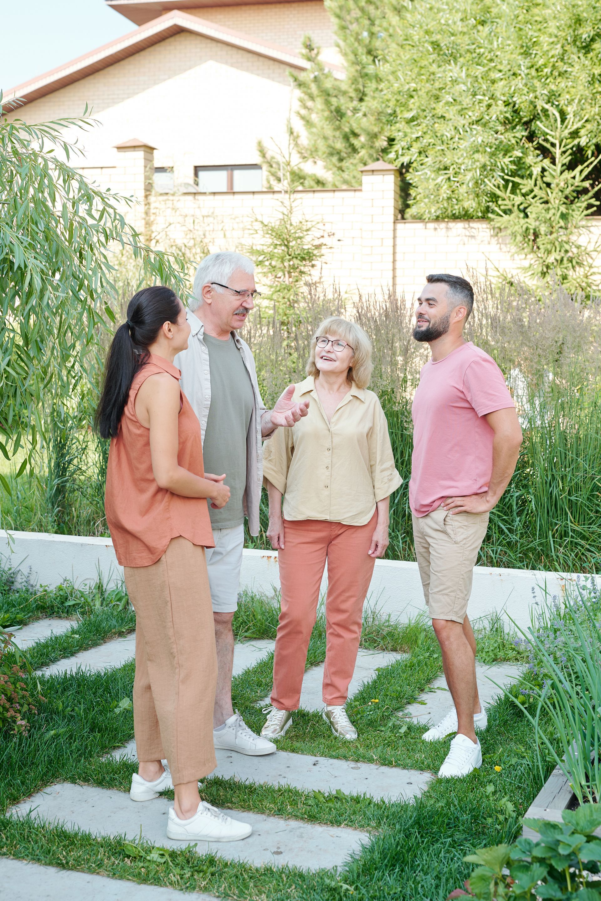 Four people chatting on a garden path beside tall greenery and a house, with stepping stones in the lawn.