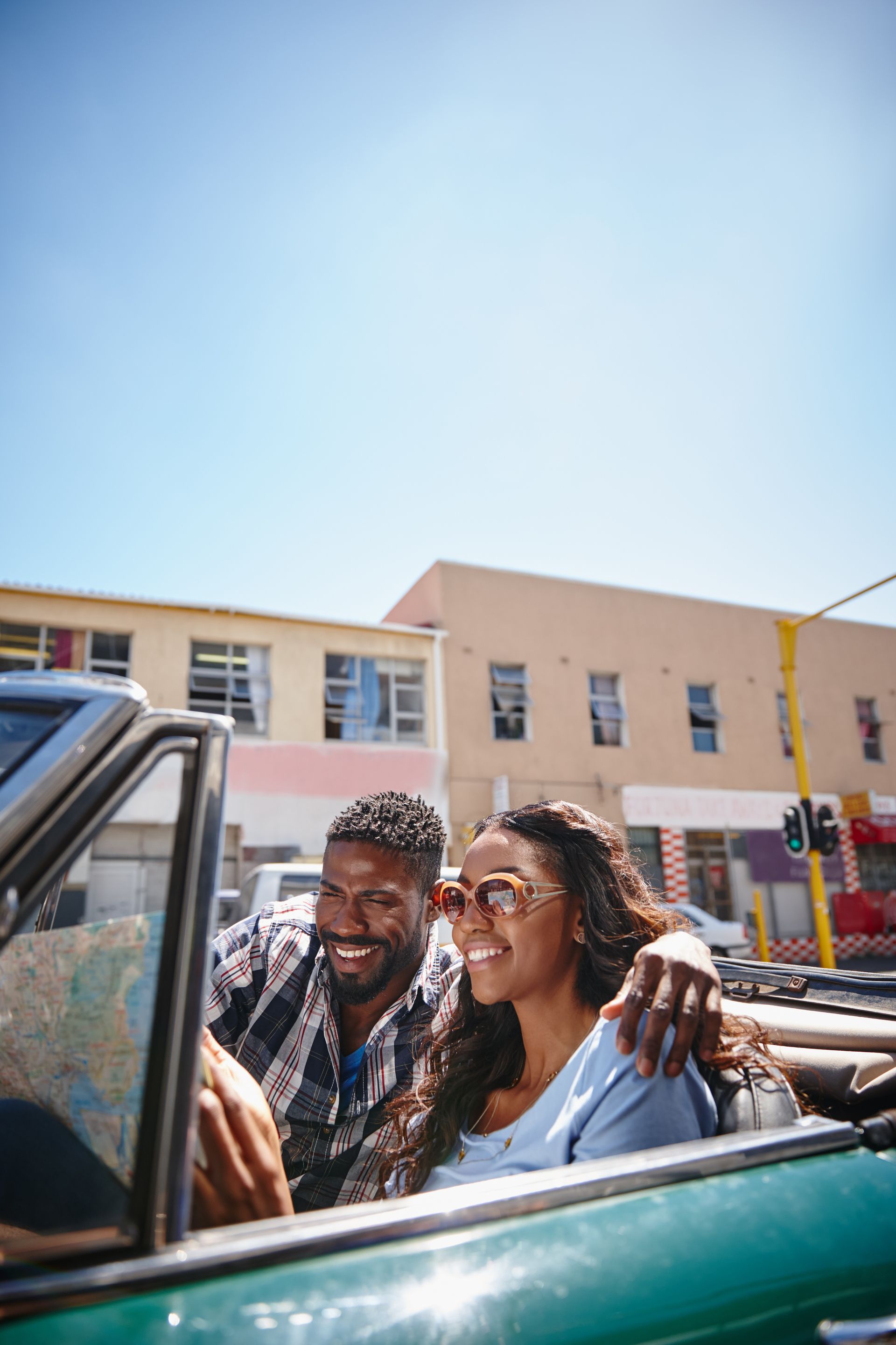 Two smiling people in a green convertible on a sunny street with pastel buildings.