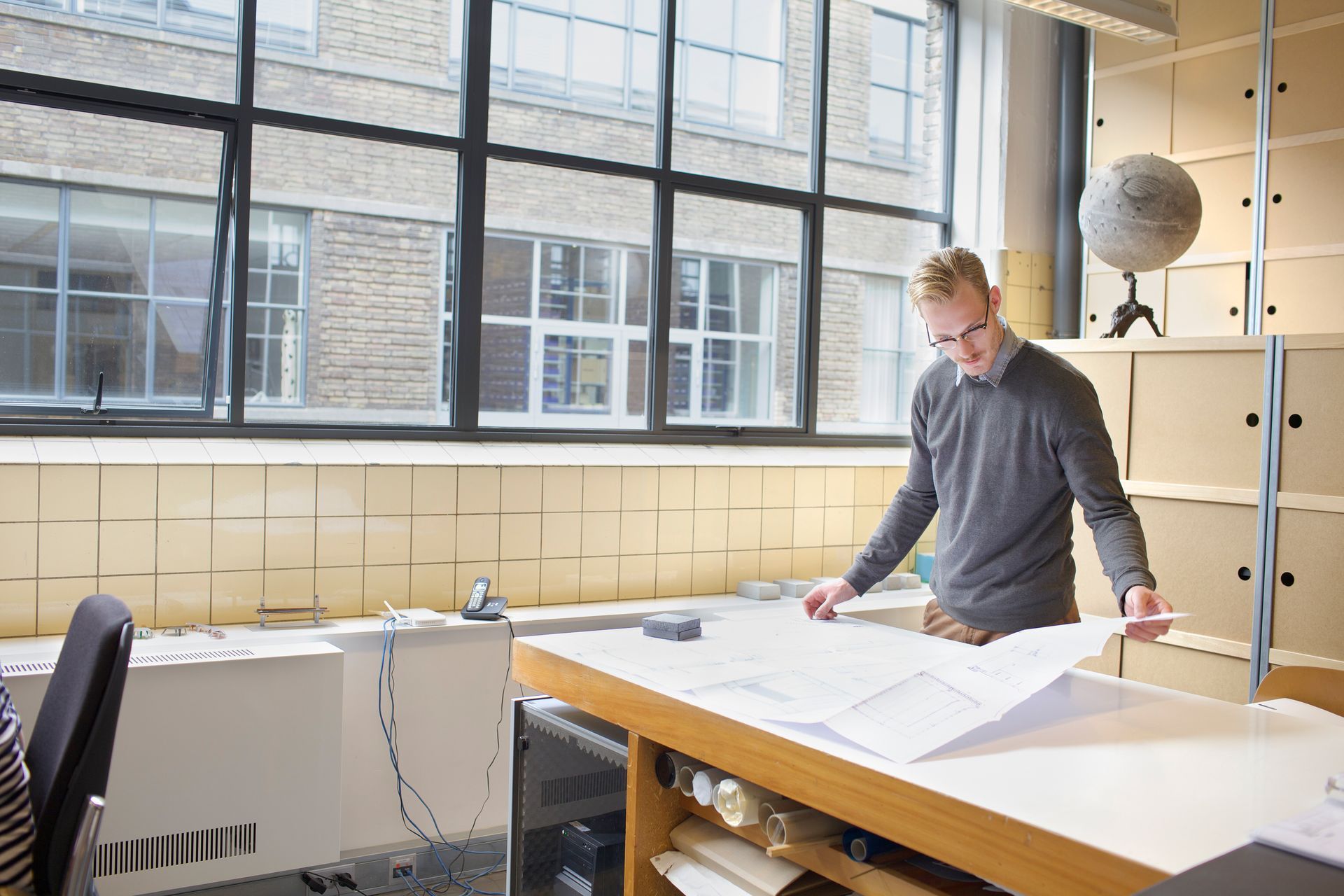 Person standing at a large drafting table in a bright studio with big windows.