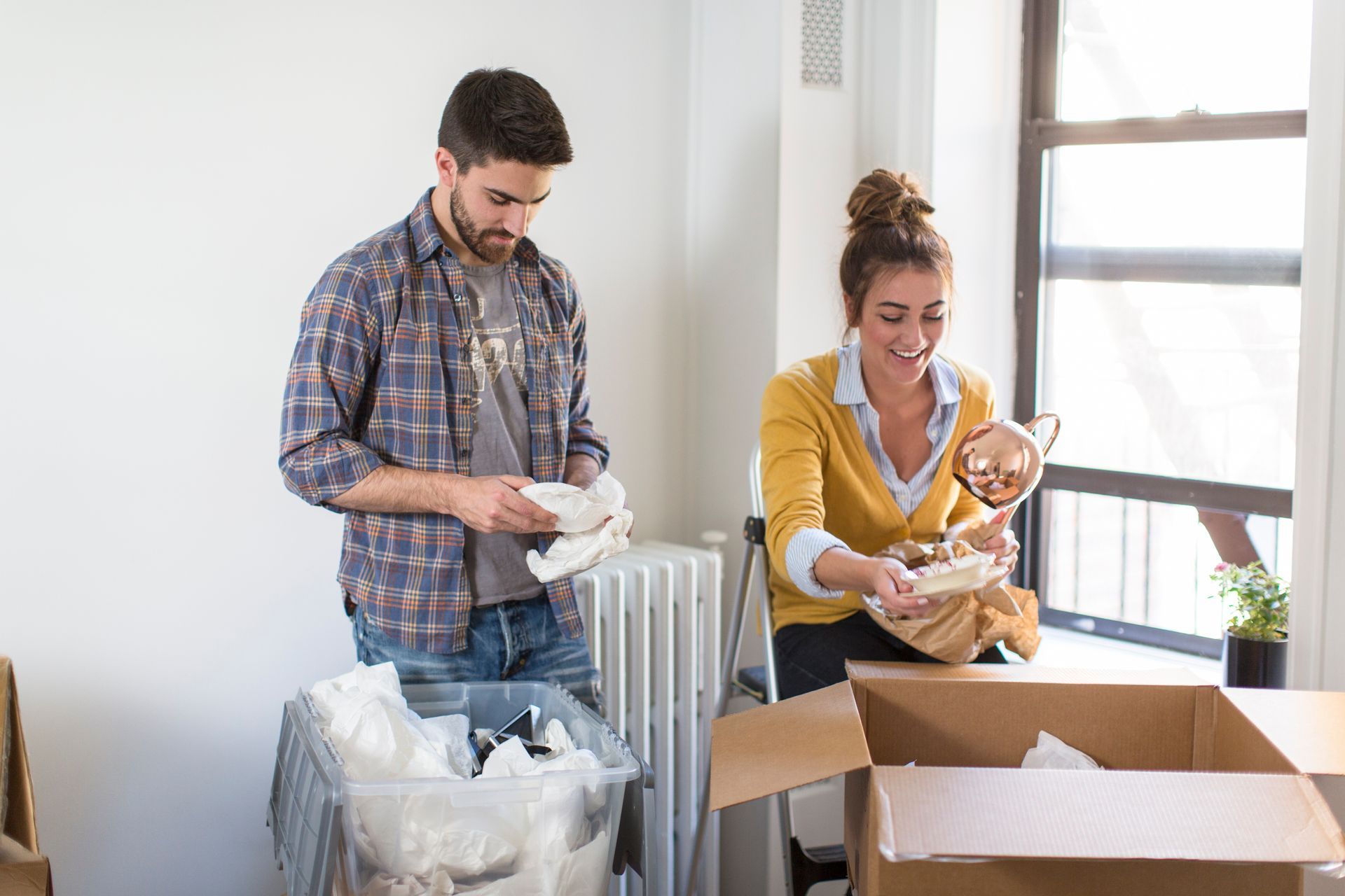 Two people packing household items into boxes in a bright room with moving supplies