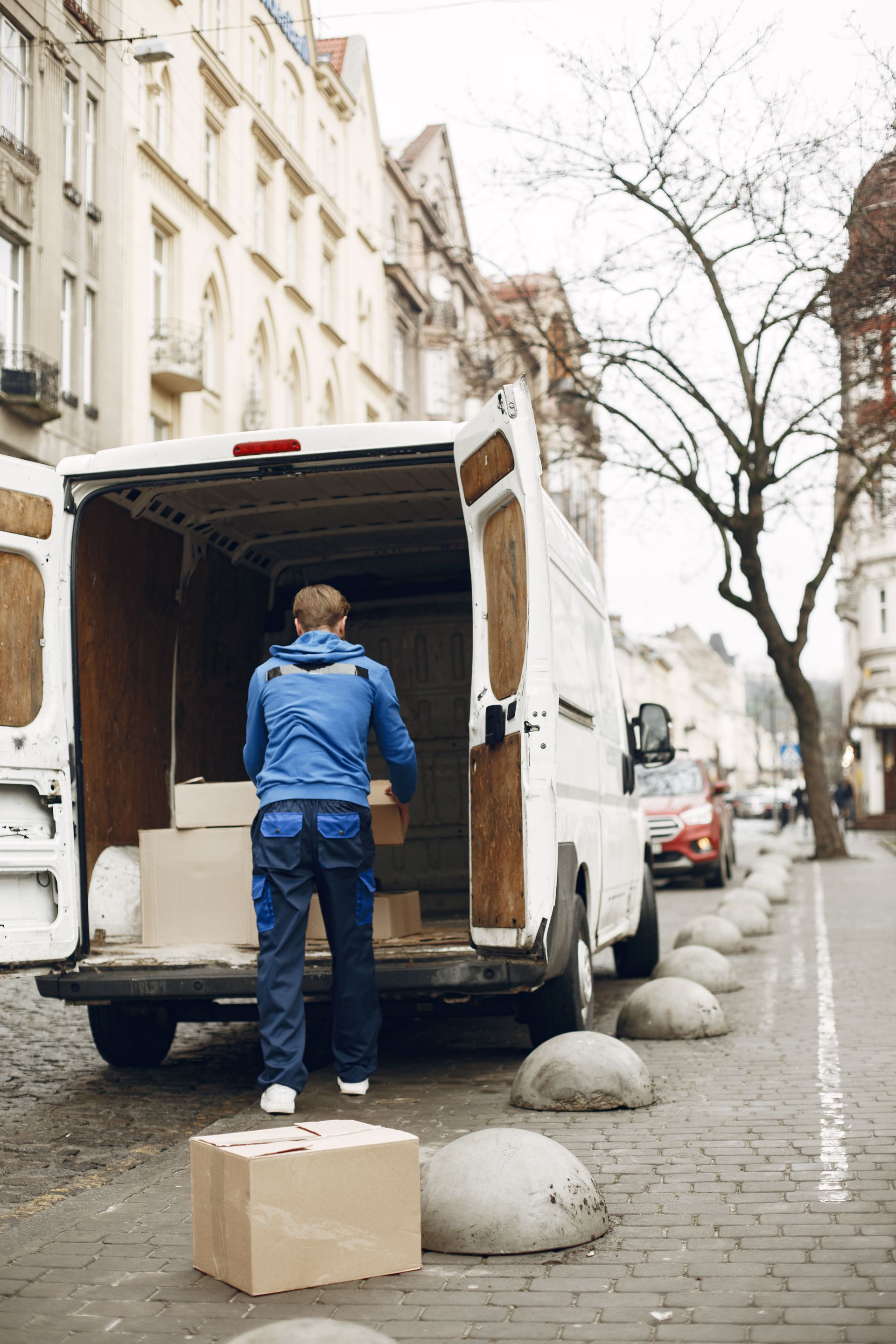 Delivery worker unloading boxes from a van on a narrow city street with parked cars and bare trees