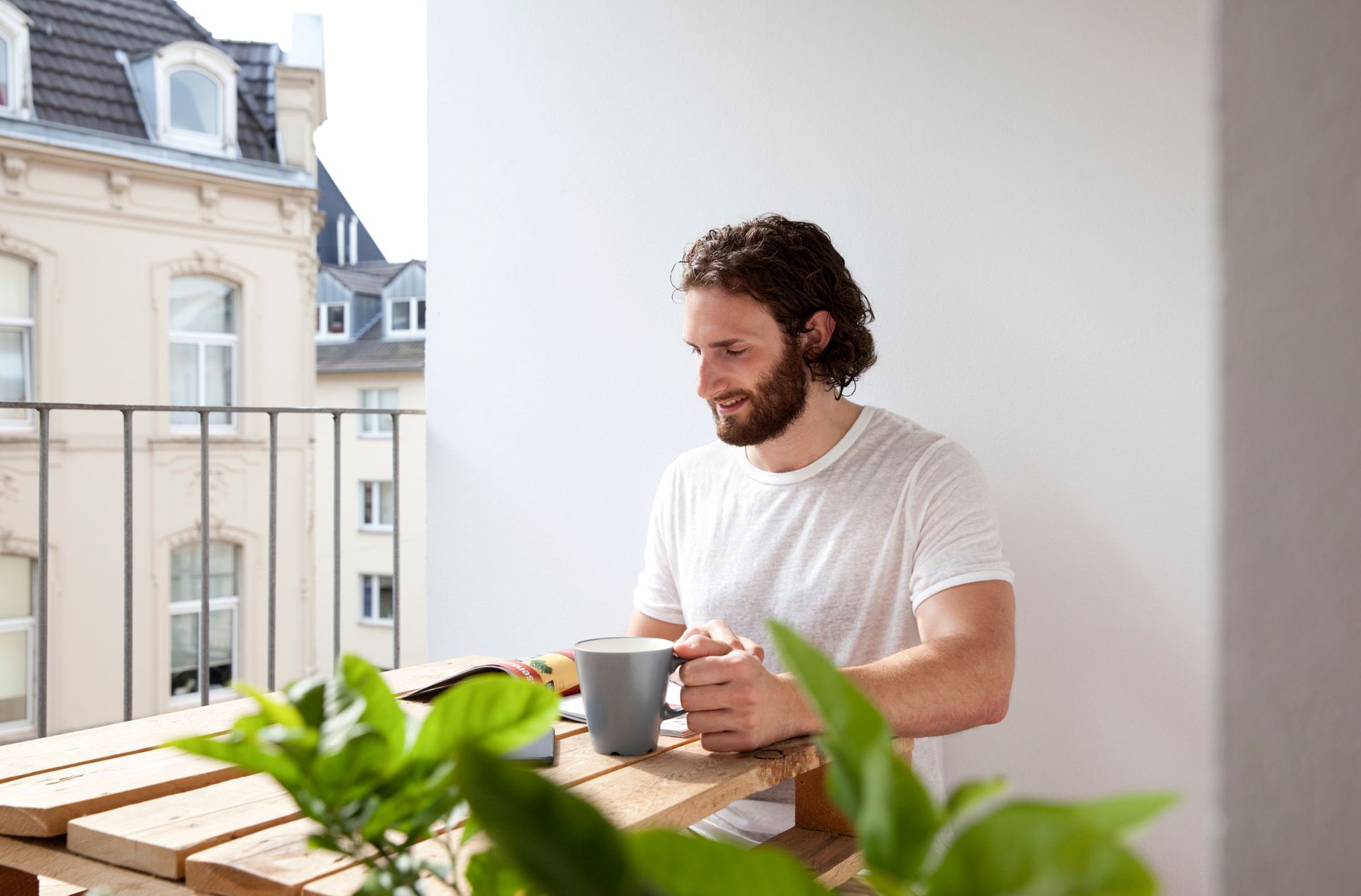 Man sipping coffee at a balcony table with a laptop, overlooking nearby apartment buildings