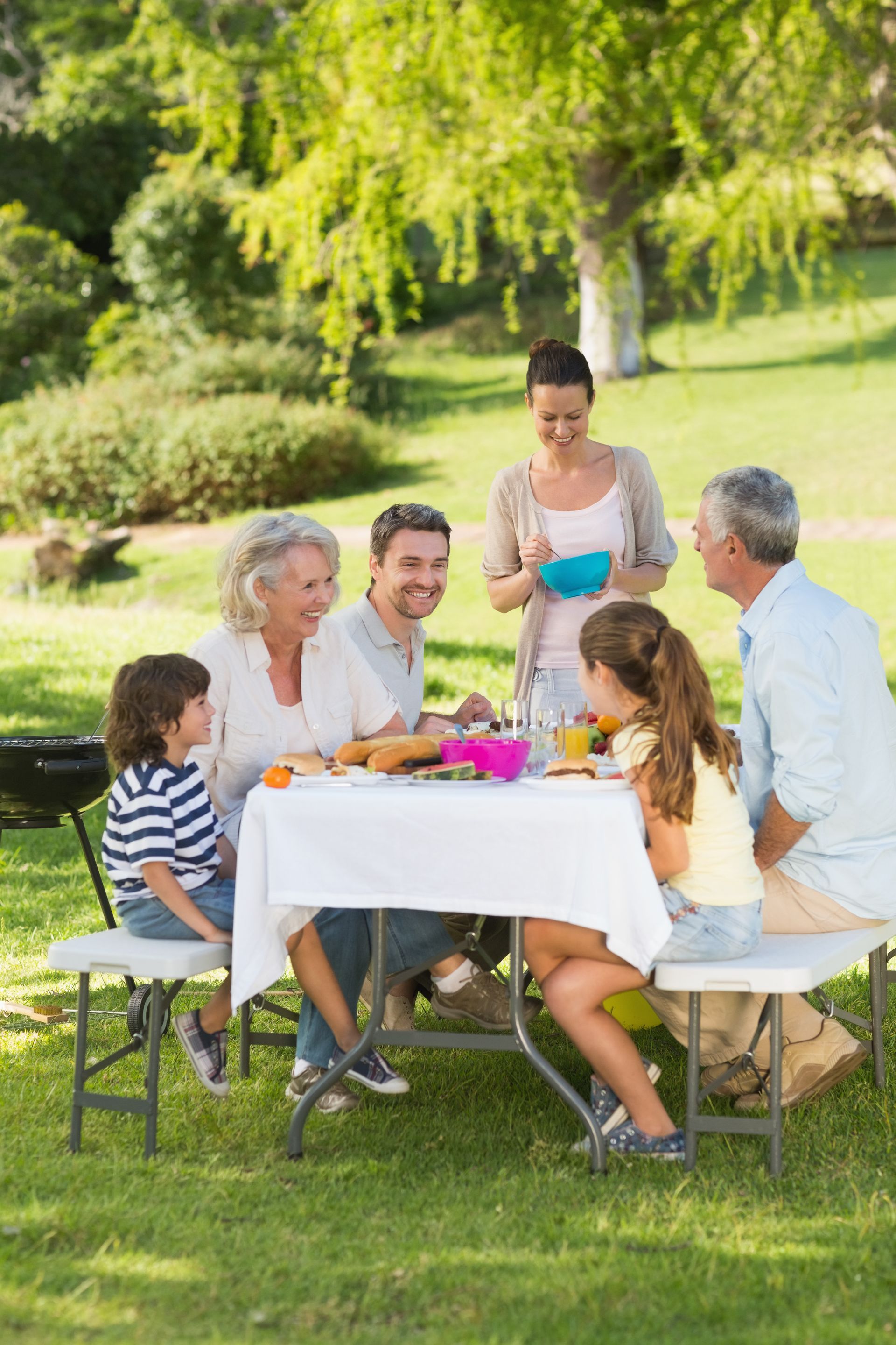 Family gathered around a picnic table in a sunny park, sharing food and drinks.