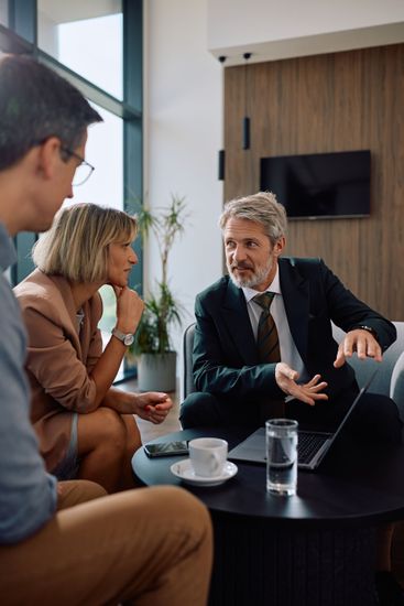 Three people in a meeting discussing a laptop at a round table in a modern office lounge.