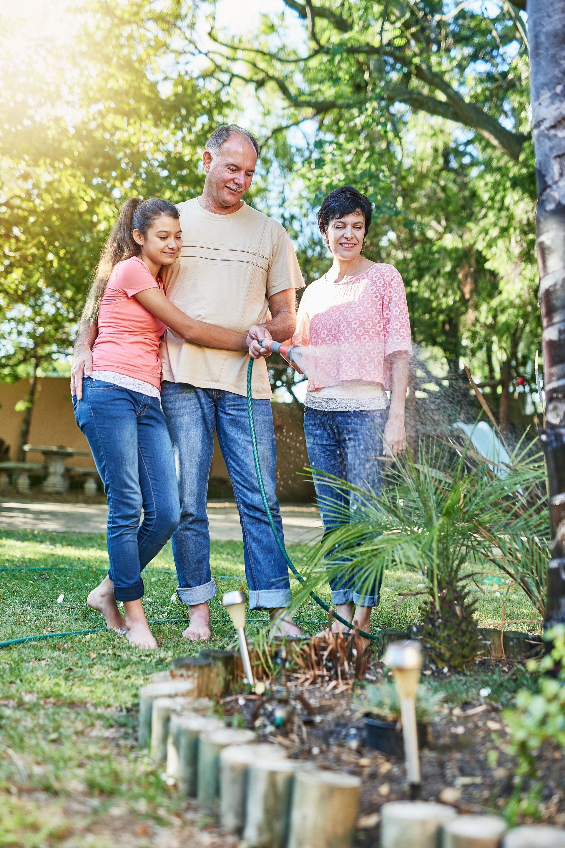Three people watering plants in a sunny garden with a hose