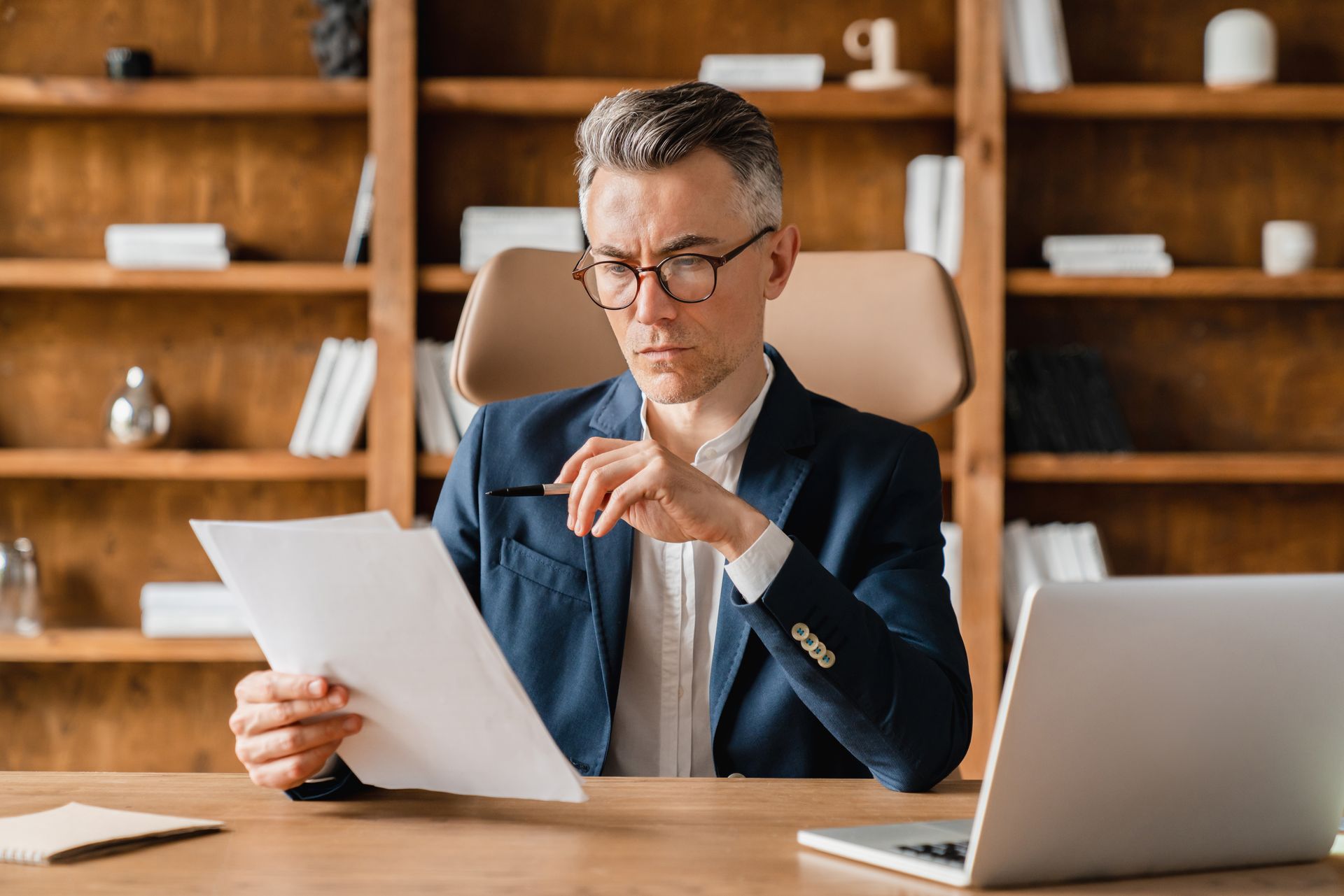 Man reading a document at a desk with a laptop in a wooden office setting