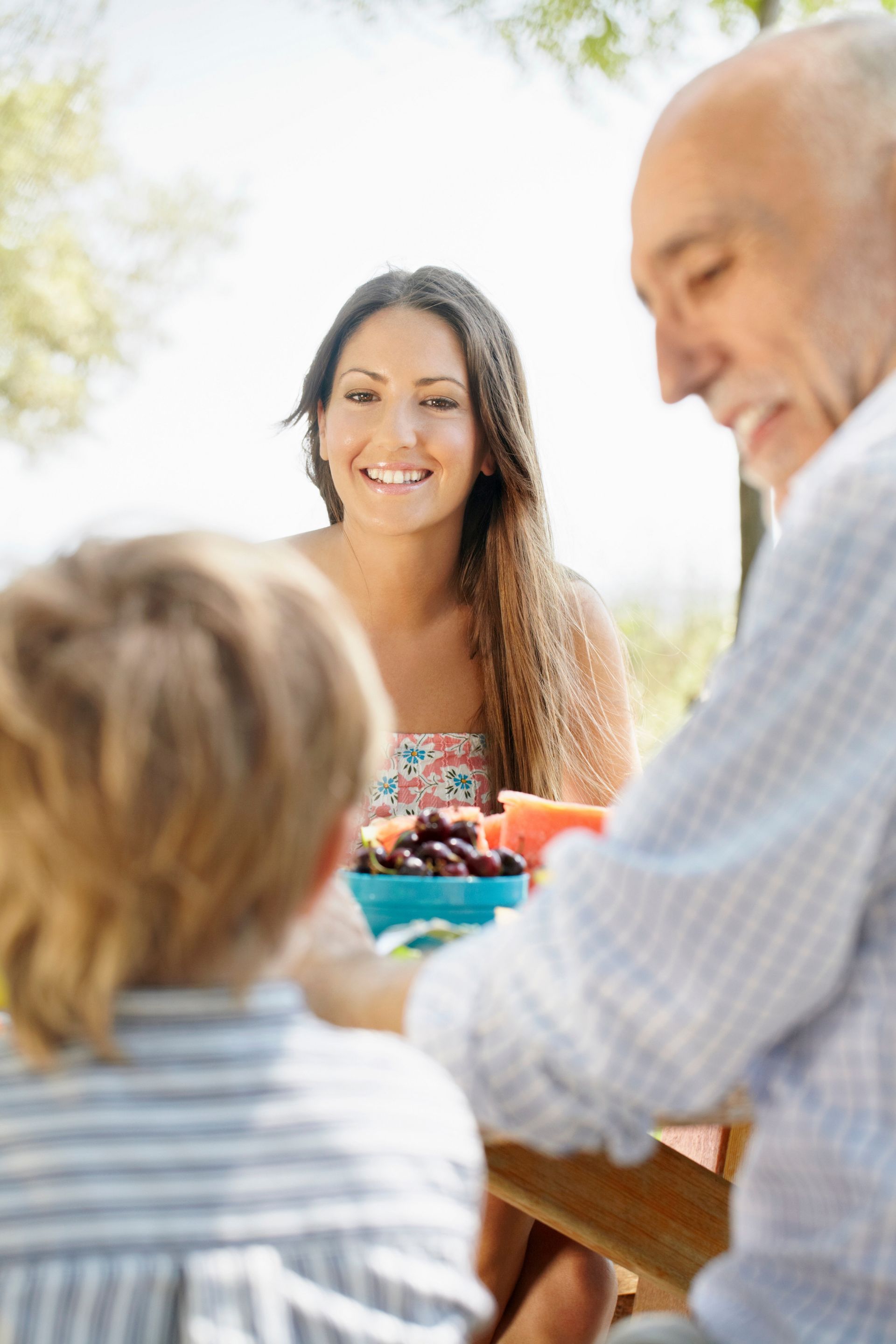 People gathered around an outdoor table, smiling and sharing a meal in bright sunlight.