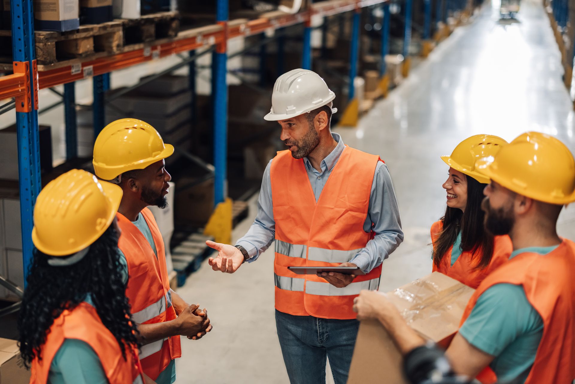Warehouse workers in hard hats and safety vests discussing beside storage racks