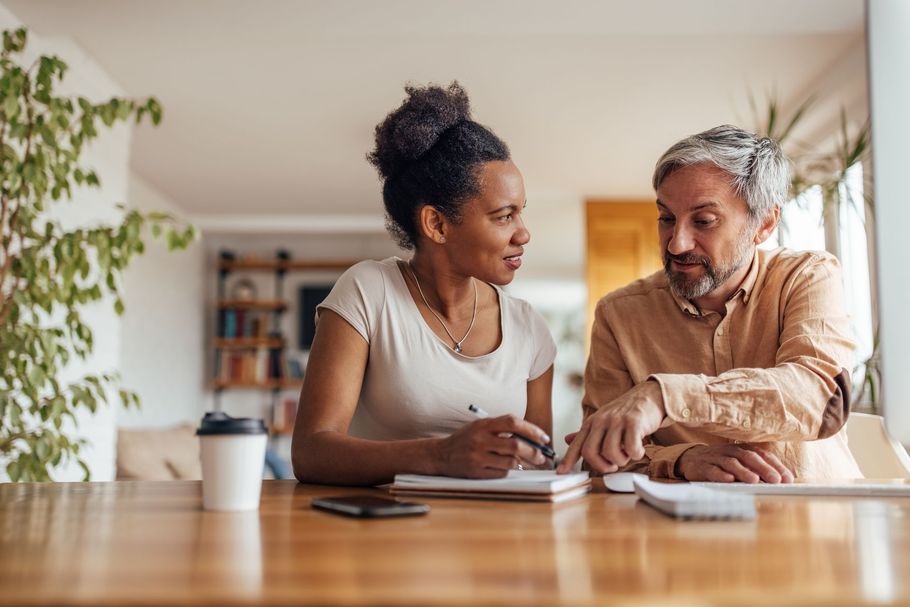Two people talking at a kitchen table, one writing in a notebook, with a coffee mug and plant nearby.