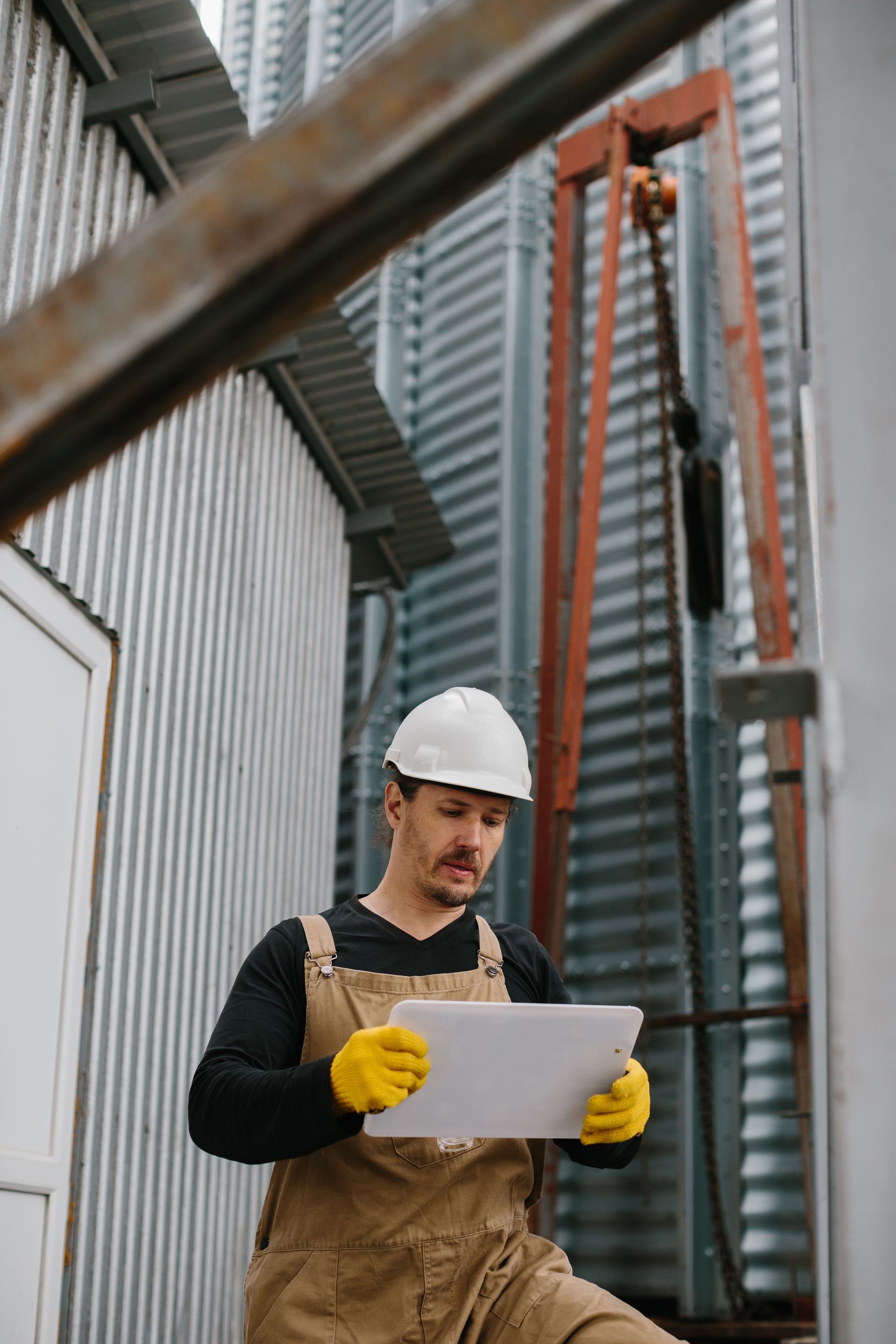 Construction worker in hard hat and gloves reviewing a clipboard at an industrial site.