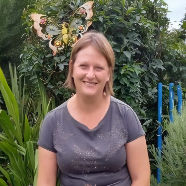 Smiling woman in a gray shirt standing in a lush garden with butterfly decorations behind her