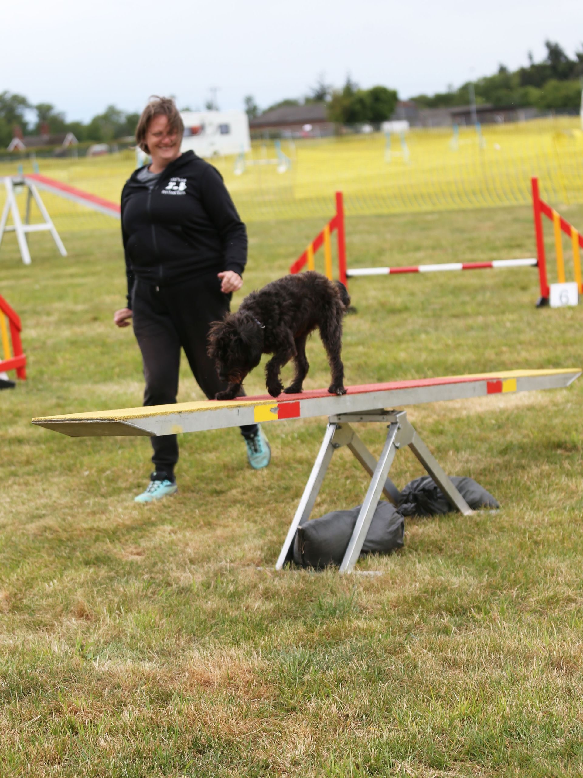 Woman stands beside a black dog balancing on a seesaw at an agility course.