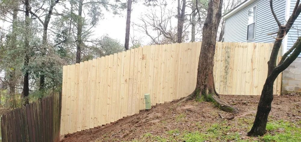 A new wooden fence on a sloped yard. Trees and a light blue house are in the background.