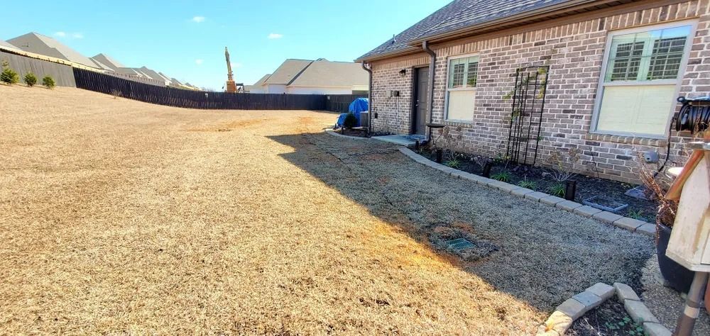 A large backyard covered in gravel with a brick house and fence. Blue sky overhead.