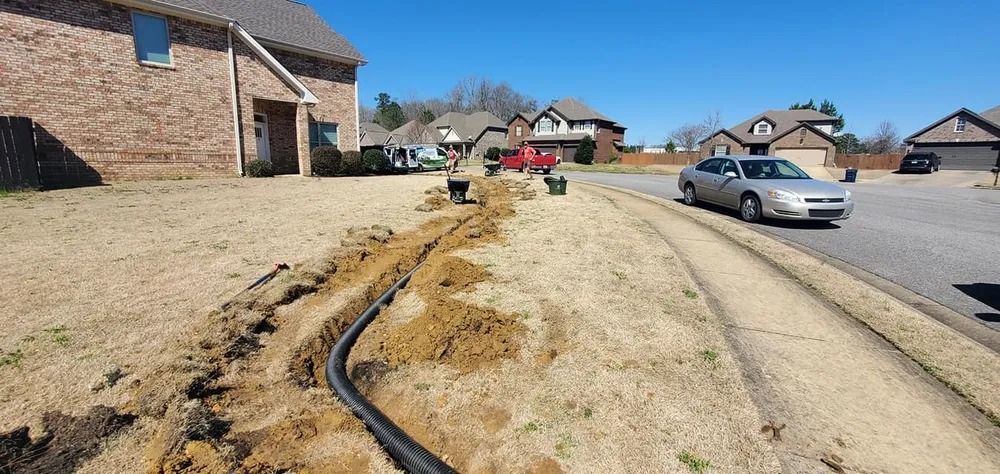 A trench dug in a yard, with black pipe, a car and houses in the background.