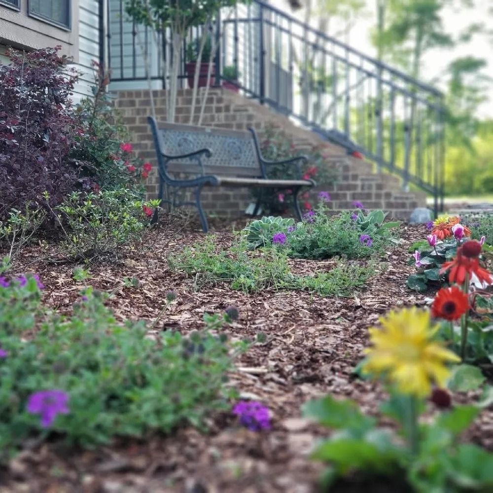 A garden bed with flowers in front of a brick staircase leading to a porch, with a bench.