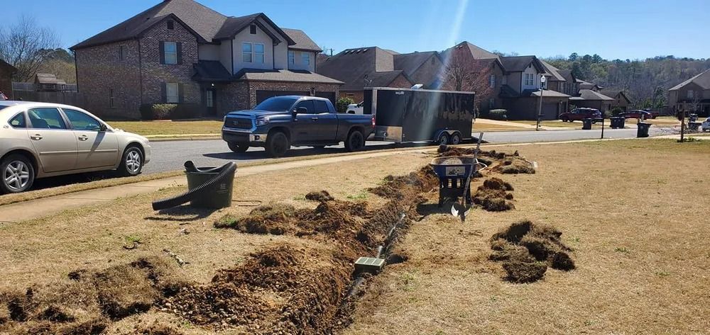 A trench dug in a residential yard; a truck and trailer parked on the street, houses in background.