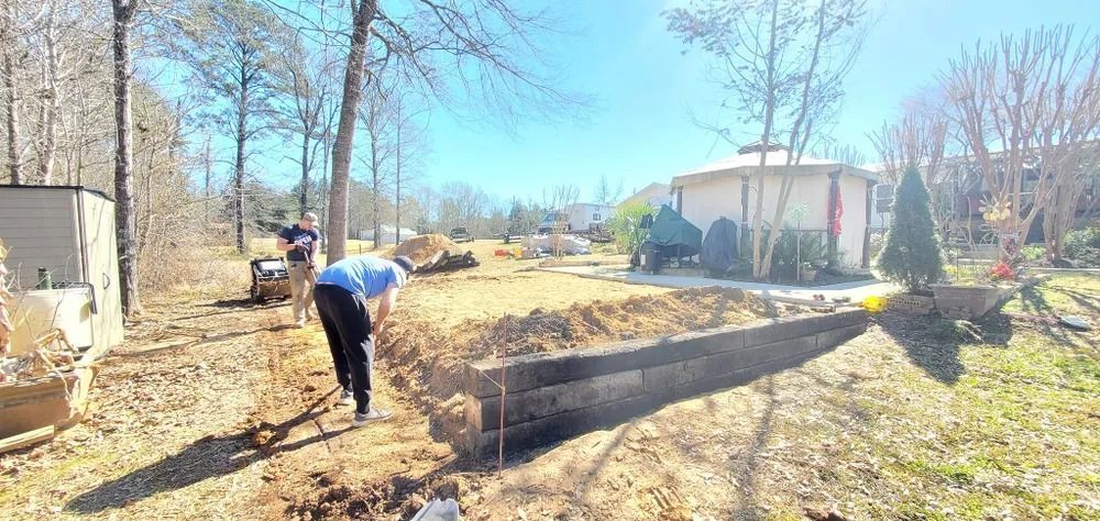 Two people working on a yard. One shovels dirt into a long trench. Sunny day. Trees and a shed are visible.