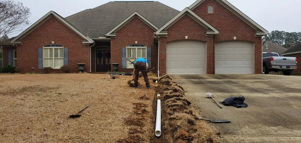 A person digging a trench in front of a brick house, laying a white pipe in the trench.