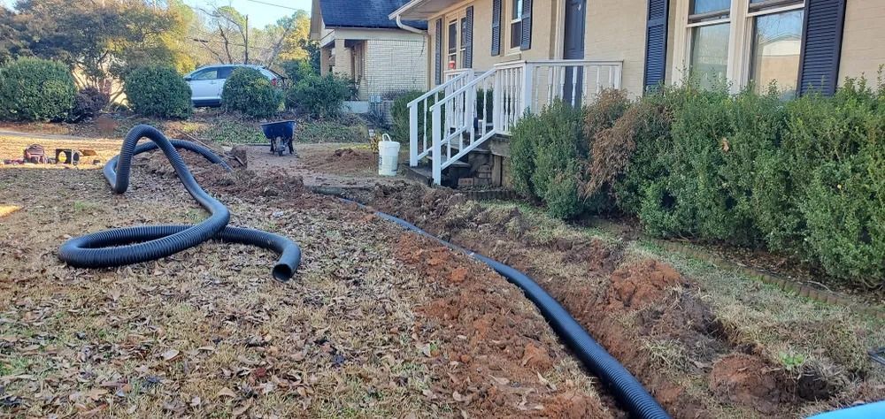 A black corrugated drainpipe runs through a dug trench in front of a house with a dog in the yard.