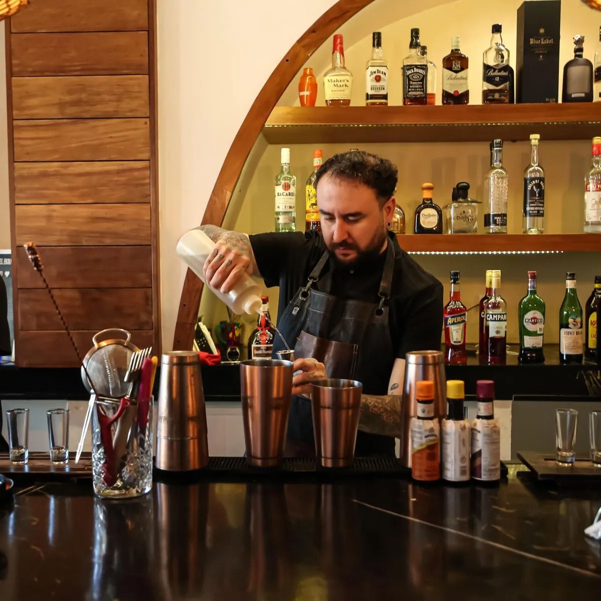 A bartender pours a white liquid from a glass bottle into a copper shaker behind a well-stocked bar.