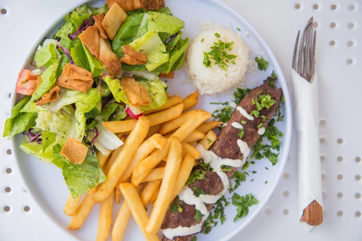 A plate featuring a grilled meat skewer with white sauce, french fries, a side salad with toasted bread, and a rice scoop.