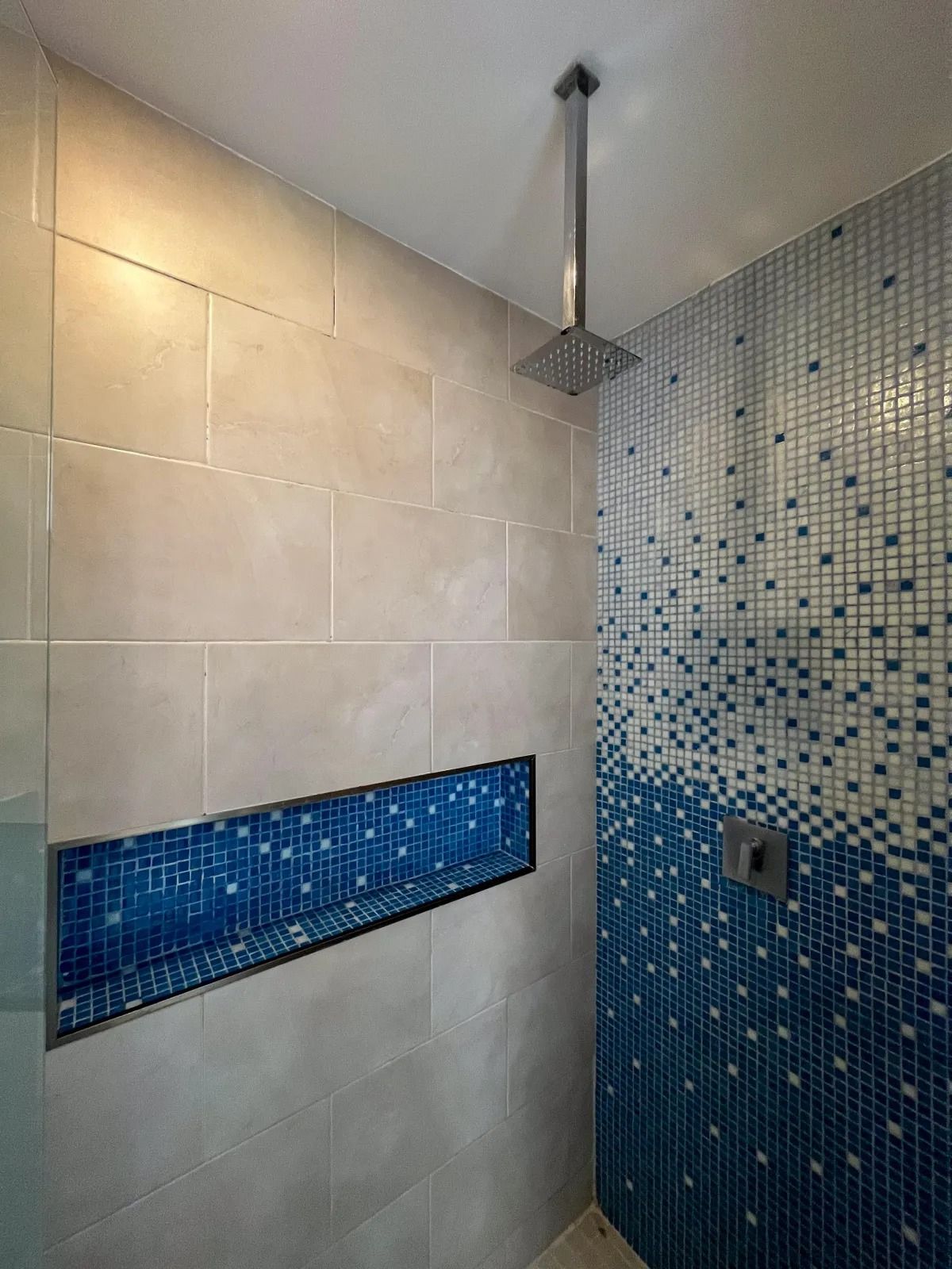 A shower with large beige tiles, a blue mosaic accent wall, a square rain showerhead, and a matching blue recessed niche.