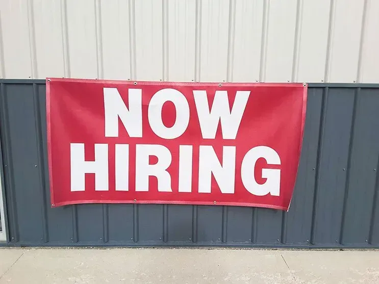 A red sign that says `` now hiring '' is hanging on a wall.
