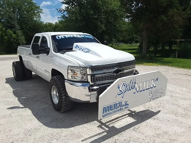A white truck is parked in a dirt lot with a sign on the front that says spit milk