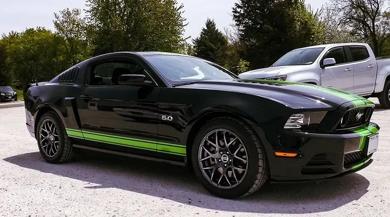 A black mustang with green stripes is parked next to a silver truck.