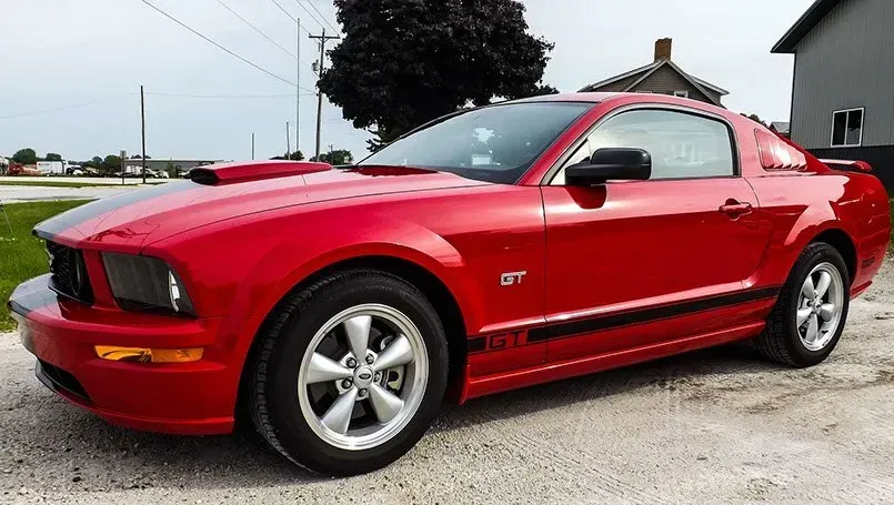 A red ford mustang is parked on gravel in front of a house.