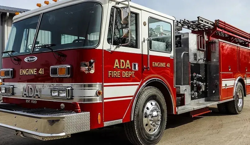 A red and white fire truck from the ada fire department is parked on the side of the road.