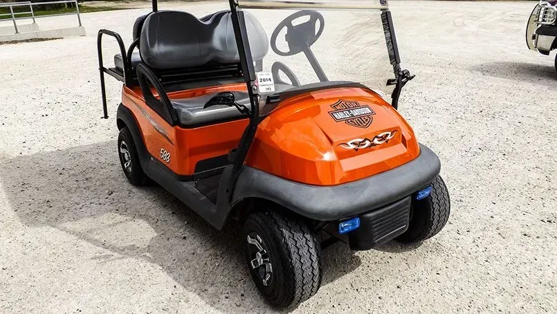 An orange golf cart is parked in a gravel lot.