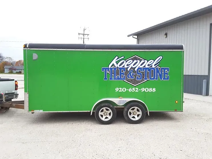 A green trailer for koeppel tile and stone is parked in front of a building.