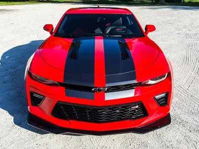 A red car with black stripes on the hood is parked on a dirt road.