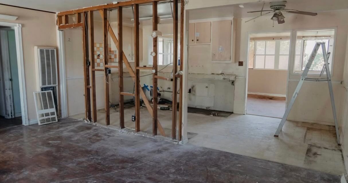 Interior during renovation; exposed wall studs, unfinished kitchen, brown floor, ladder.