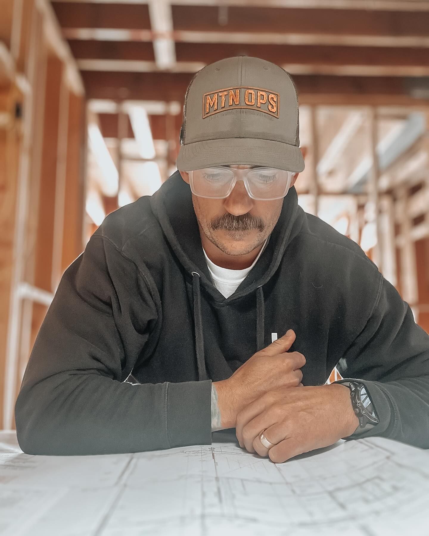 Person in a hoodie and hat with a mustache, wearing glasses, looking at blueprints in a construction site.