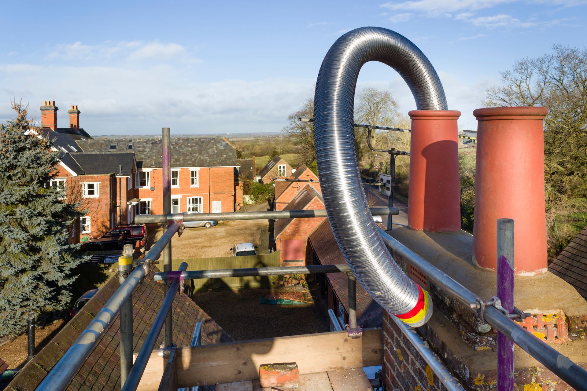 A metal flexible flue liner loops out of a brick chimney on a roof with scaffolding, set against a sunny rural landscape.