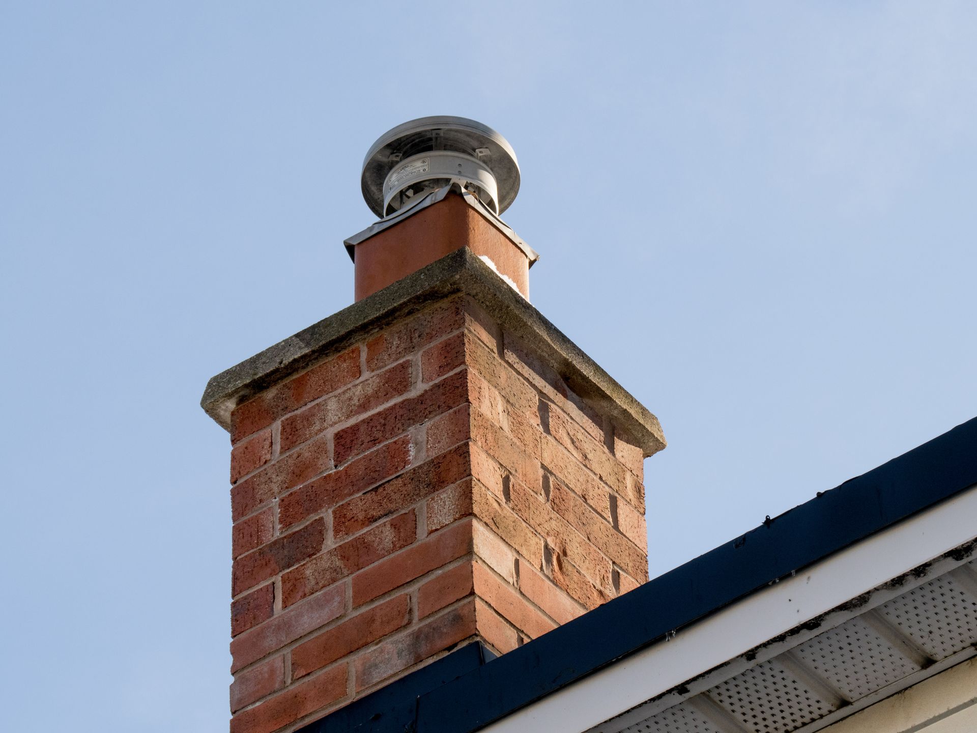 A construction worker in a high-visibility vest inspects a white chimney on a brown shingled roof under a blue sky. A construction worker in a high-visibility vest inspects a white chimney on a brown shingled roof under a blue sky.