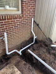 White PVC drain pipes along a brick house wall in muddy yard beside a fence