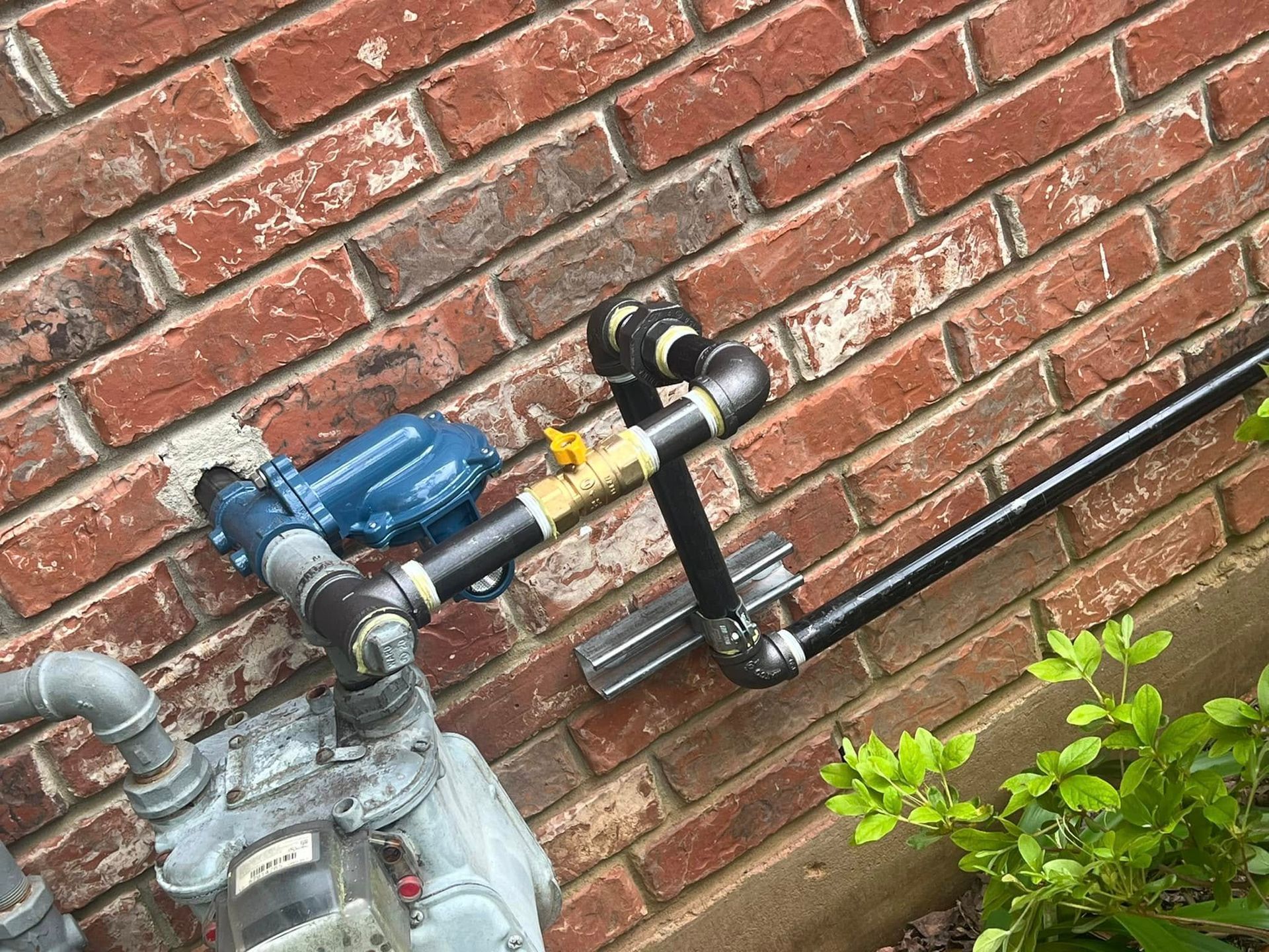 Blue gas meter and black pipe mounted on a red brick wall with a small green plant below