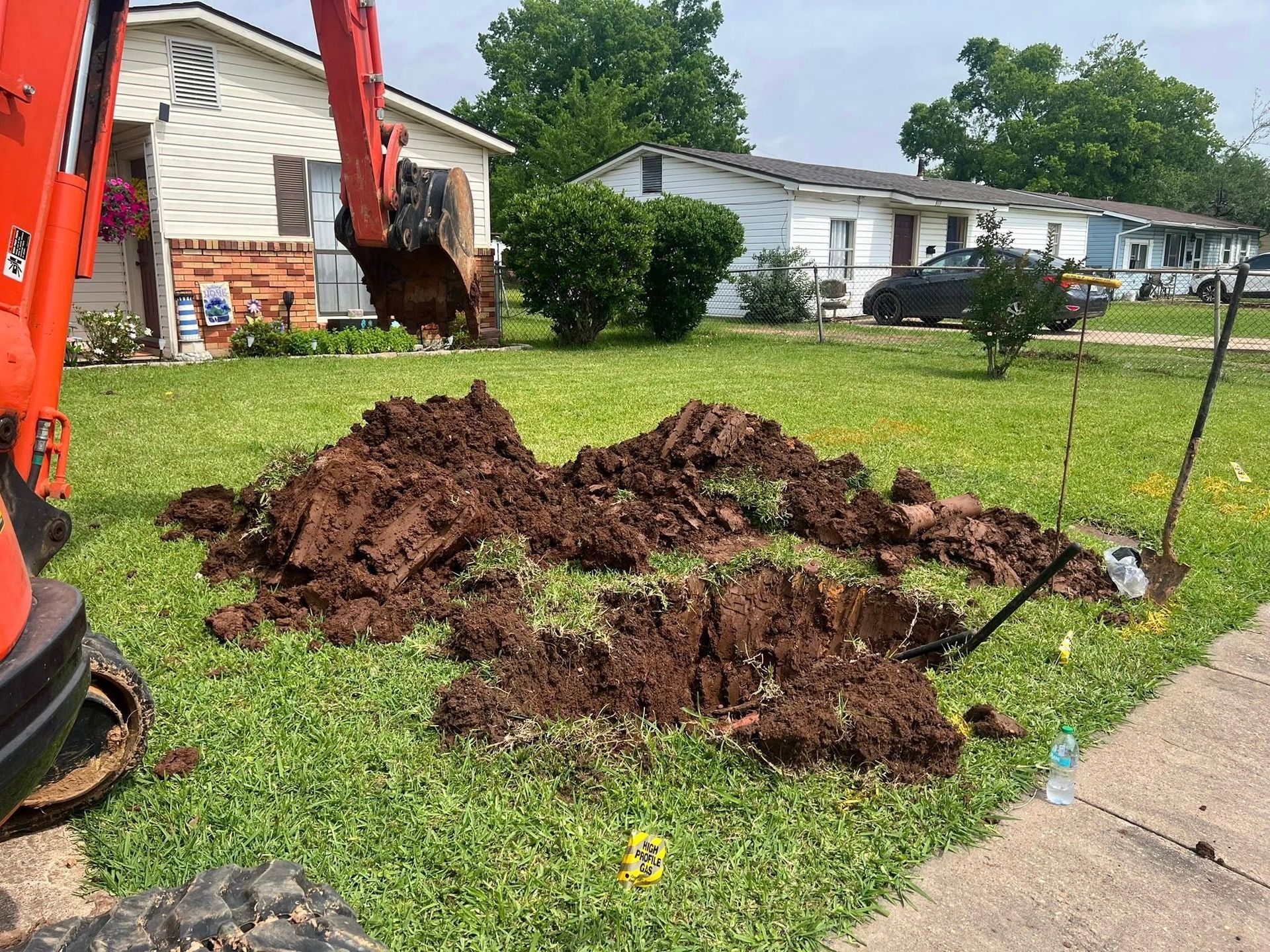 Freshly dug dirt piles beside a sidewalk in a suburban lawn, with an orange excavator arm at left.